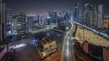 Panorama showing Bay Avenue with illuminated modern towers residential development in Business Bay aerial night , Dubai, UAE. Skyscrapers with traffic on a road near big parking lot