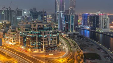 Körfez Meydanı 'nda günden geceye geçiş. Karışık kullanım ve Dubai 'deki Business Bay' de bulunan düşük katlı kompleks ofis binaları. Yukarıdan hava manzaralı ve yolda trafik var.