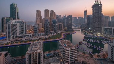 Dubai Marina with boats and yachts parked in harbor and illuminated skyscrapers around canal aerial night to day transition  before sunrise. Towers of JBR district on a background