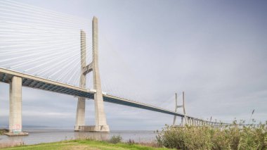 Architectural landmark Vasco da Gama Bridge over the Tagus River in Lisbon, Portugal. Green grass and cloudy sky. The longest bridge in the European Union.