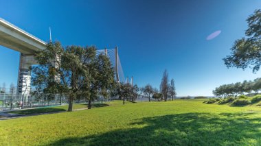 The Vasco da Gama Bridge timelapse hyperlapse viewed from green lawn with trees. Cable-stayed longest bridge flanked by viaducts and rangeviews that spans Tagus River in Park Nations. Lisbon, Portugal