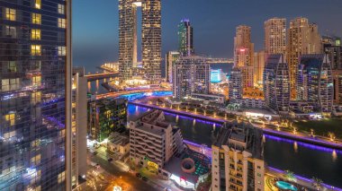 Günbatımından sonra Dubai Marina gökdelenleri ve JBR semti aydınlanmış lüks binalar ve gece geçiş zamanlarıyla dolu tatil köyleri. Palmiyeler ve kanalda yüzen teknelerle dolu bir liman.