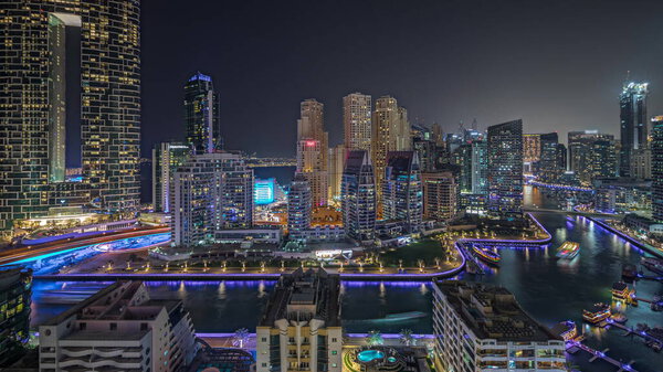 Panorama showing Dubai Marina with several boat and yachts parked in harbor and illuminated skyscrapers around canal aerial night timelapse. Towers of JBR district on a background