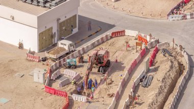 Bir grup işçi ve kırmızı kazıcı inşaat alanındaki havuzda su drenajı kazıyorlar. Yol kenarında borular tamir ediliyor.