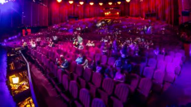 Seyirciler konser salonunda toplanıp gösteriyi zaman aşımına uğratarak izliyorlar. Kırmızı koltuklu büyük bir salon. Işığı kapatana kadar izleyiciler yerlerini dolduruyor. Bir sahneden görünüm