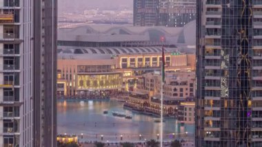 Şehir merkezindeki Dubai Çeşmesi 'nin havadan görünüşü, alışveriş merkezinin yanında palmiyeler ve bayraklar ve UAE' deki gece ve gündüz geçiş zamanının yanında. Gün batımından sonra iki gökdelen arasında görüldü.