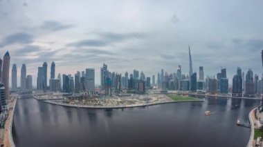 Hava görüntüsü Dubai Business Bay ve Downtown 'ın panoramik gökdelenleri ve deniz kenarı kuleleri ile kanal boyunca gündüz ve gece geçiş zamanı. Gün batımından sonra vinçleri olan inşaat alanı