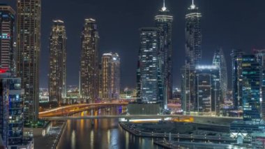 Dubai Business Bay 'in aydınlık gökdelenleri ve su kanalı hava gece zaman çizelgesine sahip şehir manzarası. Rıhtımda konut ve ofis kuleleri olan modern gökyüzü. Suda yüzen tekneler