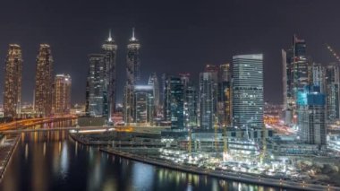 Dubai Business Bay 'in aydınlık gökdelenlerine sahip şehir manzarası su kanalı hava gece zaman çizelgesine yansıyor. Rıhtımda konut ve ofis kuleleri olan modern gökyüzü. Turnalar çalışıyor