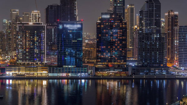 Aerial view to Dubai Business Bay and Downtown with the various skyscrapers and towers with glowing windows along waterfront on canal night timelapse. Construction site with cranes