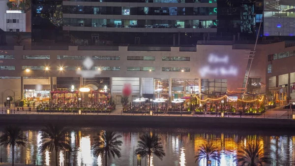 stock image Walking promenade along water canal with many crowded restaurantes in Business Bay district of Dubai aerial night timelapse. Bridges over waterfront with skyscrapers from above, United Arab Emirates