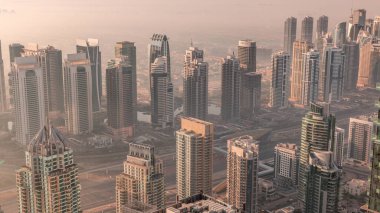 Güneş doğarken Şeyh Zayed Yolu yakınlarındaki JLT gökdelenleri ve marina kuleleri. Konut binaları ve villalı ufuk çizgisi. Sıcak turuncu ışıkla gün doğumunda sisli bir sabah
