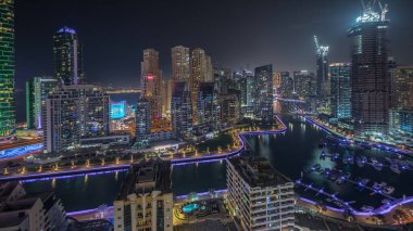Panorama, Dubai Marina gökdelenlerini ve JBR bölgesini lüks binalar ve tatil köylerinin hava geceleriyle gösteriyor. Aydınlatılmış rıhtım ve kanalda yüzen tekneler