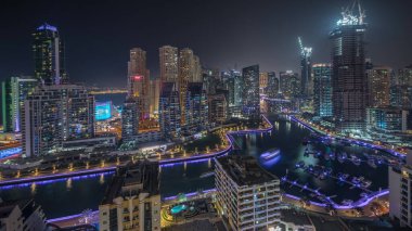 Panorama, Dubai Marina gökdelenlerini ve JBR bölgesini lüks binalar ve tatil köylerinin hava geceleriyle gösteriyor. Aydınlatılmış rıhtım ve kanalda yüzen tekneler