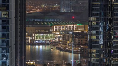 Şehir merkezindeki Dubai Çeşmesi 'nin havadan görünüşü, avuçlar ve parktaki bayraklar aydınlatılmış alışveriş merkezi ve BAE' deki Souq Night Timelapse 'ın yanında. Yukarıdan iki gökdelen arasında görüldü.