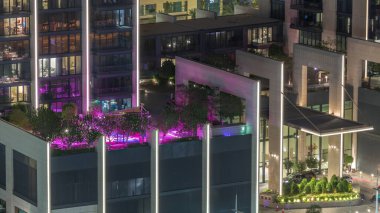 Şatafatlı yüzme havuzu, akşam havasında harika bir zaman dilimi boyunca çatıda. Palmiyeler ve ağaçlarla aydınlatılmış renkli su. Tatil ve tatil kavramı. Gökdelenlerin parlayan pencereleri. Dubai, BAE