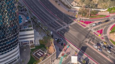 Yukarıdan Dubai zaman dilimindeki işlek yol kavşağına kadar hava görüntüsü. Renkli arabalar her iki yönde de dümdüz gidiyor ve yayalar yoldan geçiyor. Otoparkı olan finans bölgesi