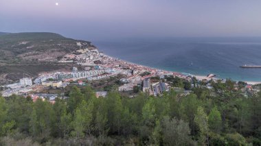 Aerial view of the coastline of the village of Sesimbra day to night transition timelapse, in the middle of the Atlantic Ocean. Evening panorama with illuminated houses from the Castle viewpoint. Portugal