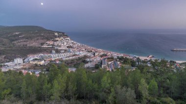 Aerial view of the coastline of the village of Sesimbra day to night transition timelapse, in the middle of the Atlantic Ocean. Evening panorama with illuminated houses from the Castle viewpoint. Portugal