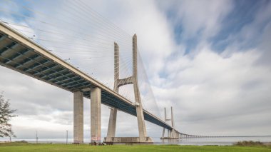 Architectural landmark Vasco da Gama Bridge over the Tagus River in Lisbon, Portugal. Green lawn with trees and cloudy sky. The longest bridge in the European Union.