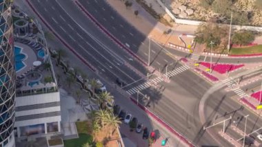 Yukarıdan Dubai zaman dilimindeki işlek yol kavşağına kadar hava görüntüsü. Renkli arabalar her iki yönde de dümdüz gidiyor ve yayalar yoldan geçiyor. Otoparkı olan finans bölgesi