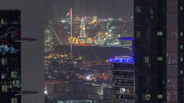 ダウンタウンの夜のタイムラプス近くにドバイ市内のアパートやヴィラに2つの高層ビルの間の空中ビュー 旗竿に旗を掲げろ 遠くに海港のライトアップされたクレーン アラブ首長国連邦 — ストック動画
