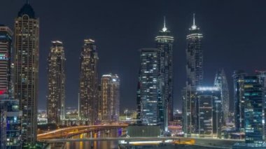 Dubai Business Bay 'in aydınlık gökdelenleri ve su kanalı hava gece zaman çizelgesine sahip şehir manzarası. Rıhtımda konut ve ofis kuleleri olan modern gökyüzü. Suda yüzen tekneler