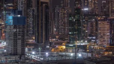 Dubai Business Bay 'in aydınlık gökdelenleri ve su kanalı hava gece zaman çizelgesine sahip şehir manzarası. Rıhtımda konut ve ofis kuleleri olan modern gökyüzü. İnşaat halindeki binalar