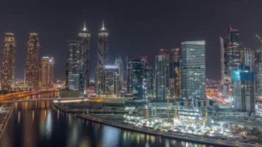 Dubai Business Bay 'in aydınlık gökdelenlerine sahip şehir manzarası su kanalı hava gece zaman çizelgesine yansıyor. Rıhtımda konut ve ofis kuleleri olan modern gökyüzü. Turnalar çalışıyor