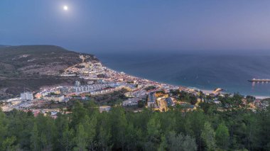 Aerial view of the coastline of the village of Sesimbra day to night transition timelapse, in the middle of the Atlantic Ocean. Evening panorama with illuminated houses from the Castle viewpoint. Portugal