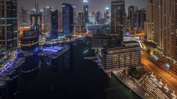 Aerial panoramic view to Dubai marina illuminated skyscrapers around canal with floating yachts timelapse during all night and lights turning off. White boats are parked in yacht club