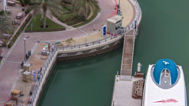 İnşaatçılar, Dubai Marina 'da su nakliye istasyonu anteninin yakınındaki palmiyelerle rıhtımın yeniden inşasını yapıyorlar. Yeni sarı tuğlalı fayanslar kaldırımda. Yatlar ve tekneler suda yüzüyor.