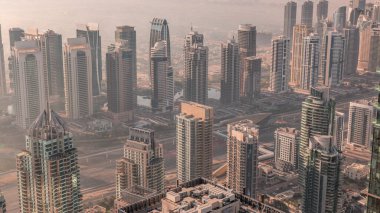 Güneş doğarken Şeyh Zayed Yolu yakınlarındaki JLT gökdelenleri ve marina kuleleri. Konut binaları ve villalı ufuk çizgisi. Sıcak turuncu ışıkla gün doğumunda sisli bir sabah