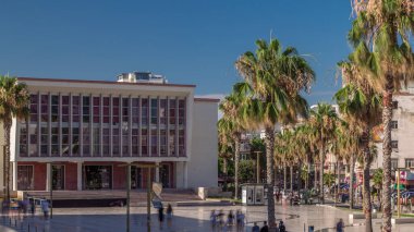Arnavutluk 'un Durres kentindeki ana meydandaki çeşme ve palmiye manzarası (Sheshi Liria) ve tiyatro önünde yürüyüş alanı bulunuyor. Sokakta trafik vardı ve insanlar dolaşıyordu.