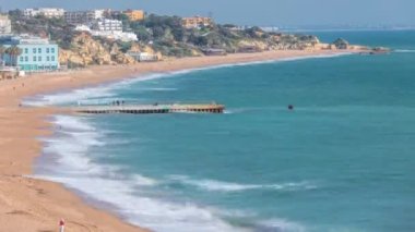 Geniş kumlu sahil ve Albufeira zamanındaki Atlantik Okyanusu. Kayalıkların tepesindeki beyaz ve turuncu evler. İskelede birileri var. Yukarıdan hava görüntüsü. Algarve, Portekiz