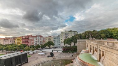 Panorama, Lizbon 'daki Jardim da Alameda' nın gün batımında Aydınlık Çeşme (Fonte Luminosa) zaman ayarlı görüntüsünü gösteriyor. Meydanı çevreleyen yeşil çimenli renkli evlerin üzerinde dramatik bulutlar