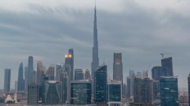 Dubai Business Bay ve Downtown 'ın havadan görüntüsü. Kanal boyunca çeşitli gökdelenler ve kuleler var. Gün ve gece geçiş zamanlarında. Gün batımından sonra vinçleri olan bir inşaat alanı. Bulutlu