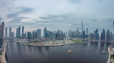 Hava görüntüsü Dubai Business Bay ve Downtown 'ın panoramik gökdelenleri ve deniz kenarı kuleleri ile kanal boyunca gündüz ve gece geçiş zamanı. Gün batımından sonra vinçleri olan inşaat alanı