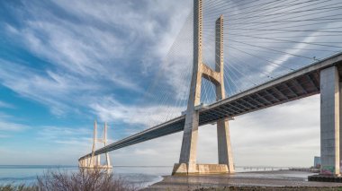 Vasco da Gama bridge timelapse hyperlapse with reflection on water and blue cloudy sky. Cable-stayed bridge and Tagus river. Lisbon, Portugal.