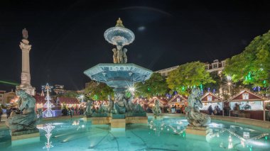 Portekiz, Lizbon 'un Baixa bölgesindeki Dom Pedro IV Meydanı' ndaki Rossio Noel Pazarı 'nda akşam süslemeleri ve yemek kabinleriyle aydınlatılmış çeşmeyi gösteren panorama..