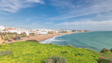 Panorama Albufeira Timelapse şehrinde geniş kumlu sahil ve Atlantik okyanusunu gösteriyor. Uçurumun tepesindeki beyaz evler. Yeşil çimlerin üzerinde sarı çiçeklerle yukarıdan hava manzarası. Algarve, Portekiz