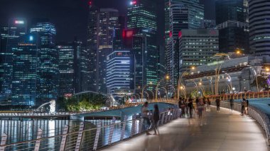 Yürüyen insanlarla Esplanade Köprüsü ve arka planda merkez gökdelenleri olan Singapur gece zaman çizelgesi. Aydınlatılmış kuleler suya yansıyor
