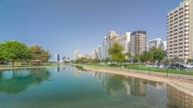 Abu Dabi 'nin sahil şeridi boyunca uzanan Corniche Bulvarı plajı parkında arka planda gökdelenler var. Lake Park 'tan yansıtmalı görüntü. Bir yanda ağaçlar