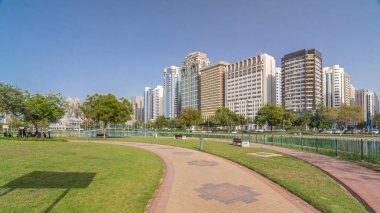 Abu Dabi 'nin sahil şeridi boyunca uzanan Corniche Bulvarı plajı parkında arka planda gökdelenler var. Lake Park 'ın manzarası. Bir tarafta ağaçlar. Güneşli bir günde mavi gökyüzü