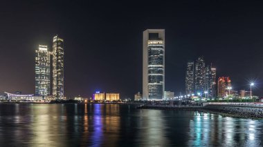 Abu Dabi Skyline 'ın panoramik görüntüsü ve gece vakti deniz manzarası, Birleşik Arap Emirlikleri. Aydınlatılmış gökdelenleri olan Corniche manzarası suya yansıyor