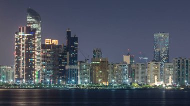 Abu Dabi Skyline 'ın panoramik görüntüsü ve gece vakti deniz manzarası, Birleşik Arap Emirlikleri. Aydınlatılmış gökdelenleri ve camii olan Corniche manzarası