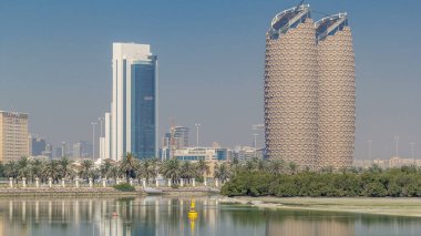 Abu Dabi zaman dilimindeki Al Bahr kuleleriyle gökdelenlerin görüntüsü. Sudaki yansımalar ve sokaktaki palmiyeler. Birleşik Arap Emirlikleri
