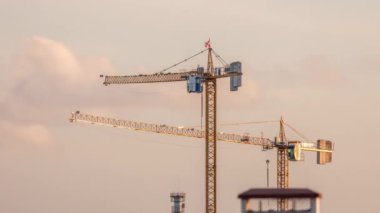 Gün batımında altından gökyüzü olan bir bina kompleksi üzerinde çalışan birkaç vinç de dahil olmak üzere büyük bir inşaat sahası. Bulutlar geçip gidiyor