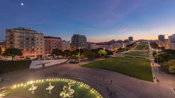 Panorama showing lawn at Alameda Dom Afonso Henriques with colorful buildings and illuminated Luminous Fountain aerial day to night transition timelapse from above after sunset in Lisbon, Portugal