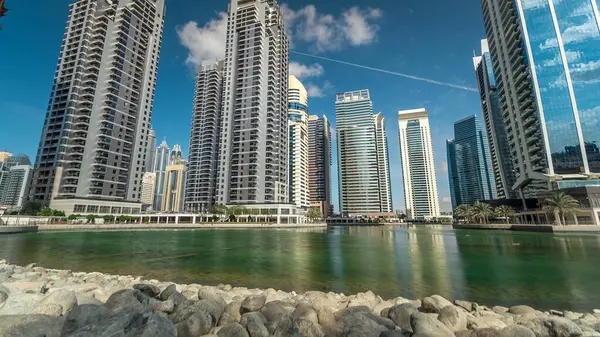 Residential buildings in Jumeirah Lake Towers reflected in water timelapse in Dubai, UAE. View with blue cloudy sky from stones near lake